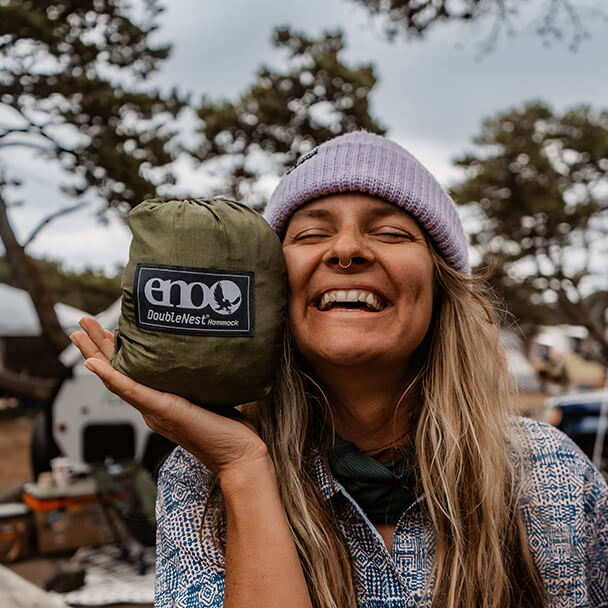 A young woman smiles with her eye closed while holding a packed ENO hammock to their cheek.