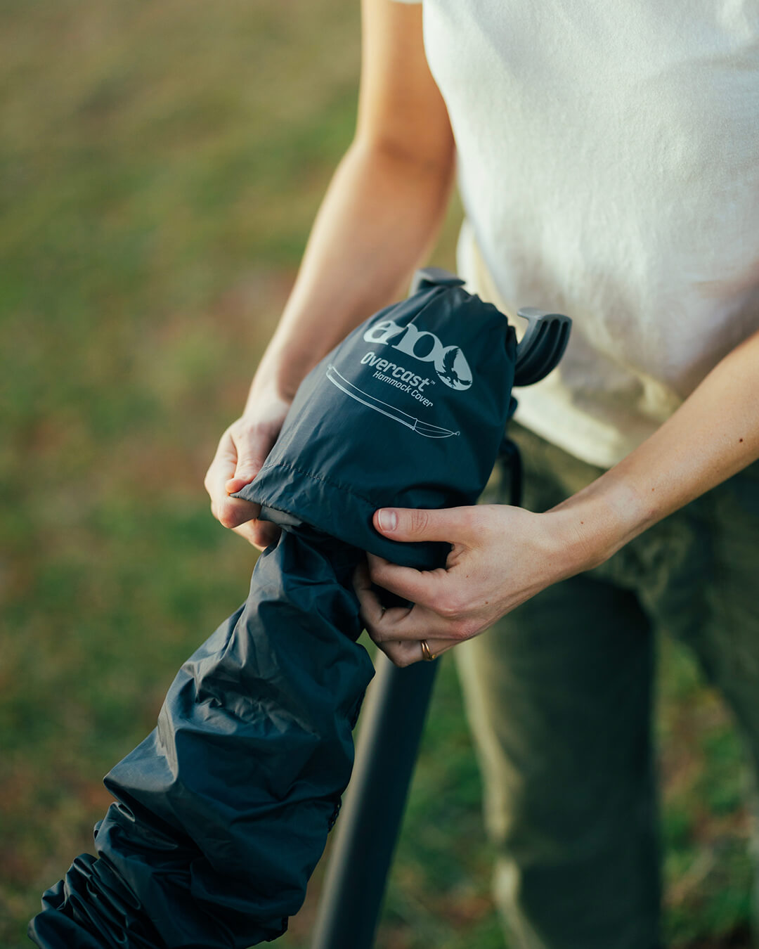 A person setting up the Overcast Hammock Cover on a hammock stand. The background shows a soft-focus grassy field.
