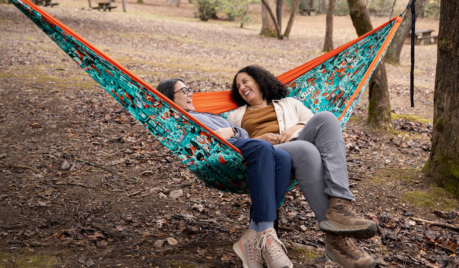 Two people sitting in the colorful Outdoor Foundation ENO DoubleNest Giving Back Hammock in a forest setting.