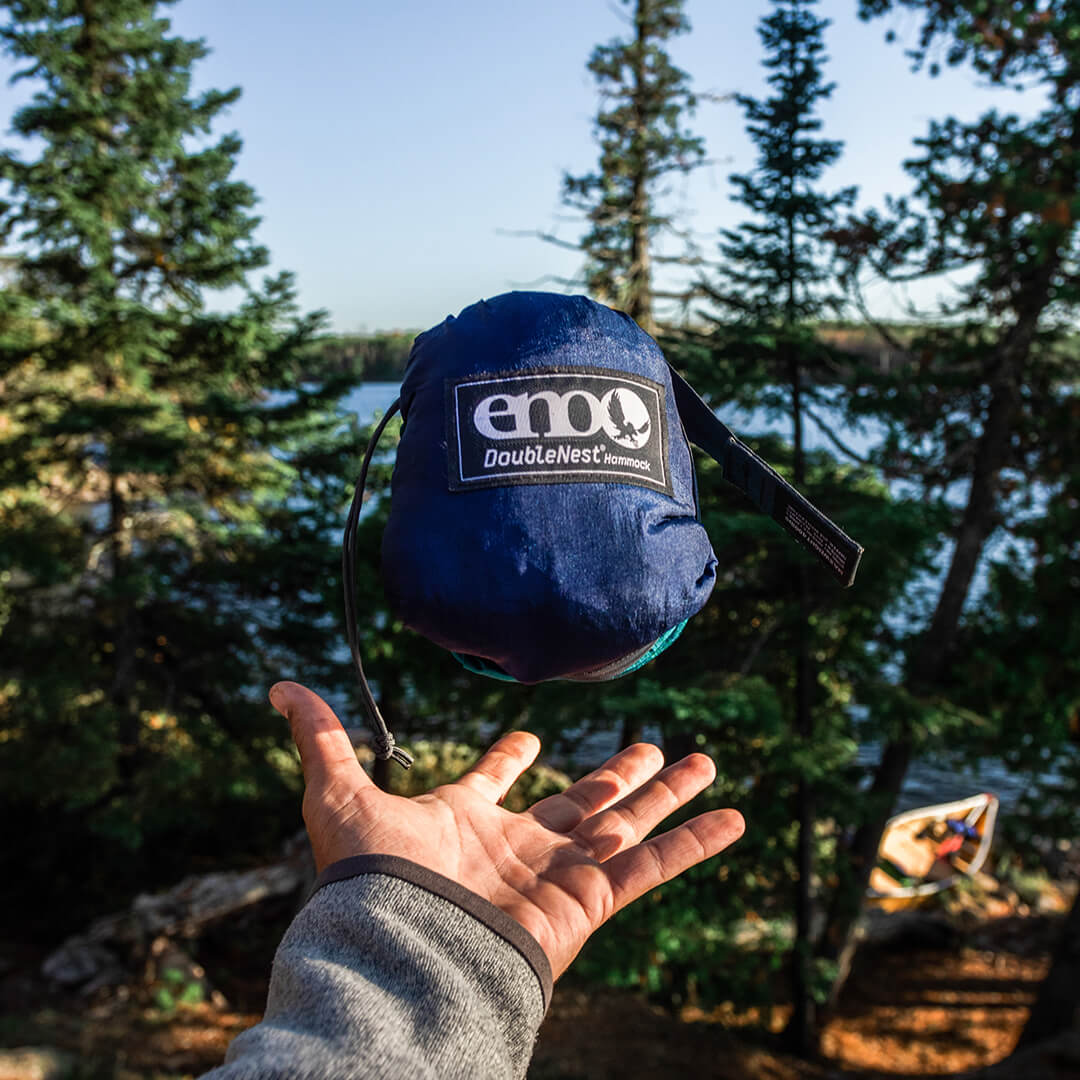 A person tosses a packed ENO portable hammock stuff sack into the air while their hand stays below ready to catch in front of a wooded lake background.