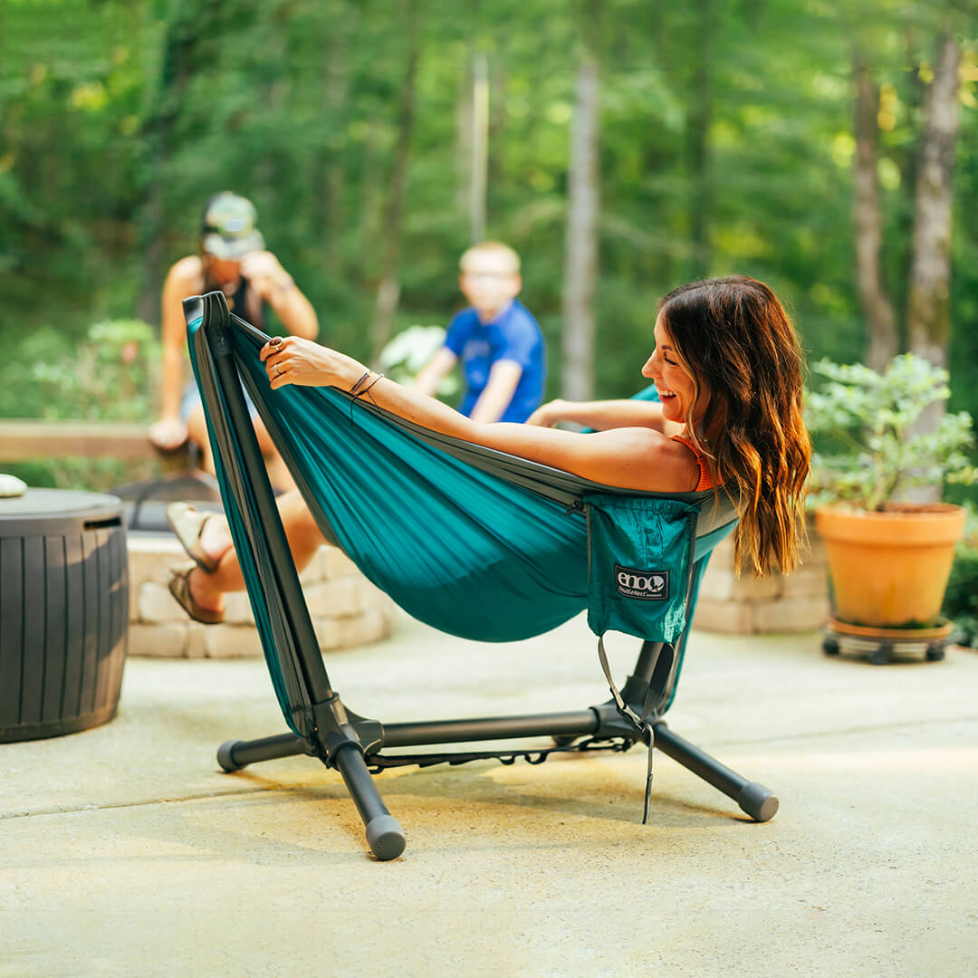 Woman smiles while leaning back in an ENO DoubleNest Hammock set up on the ENO Parklite Hammock Chair Stand with family in the background.