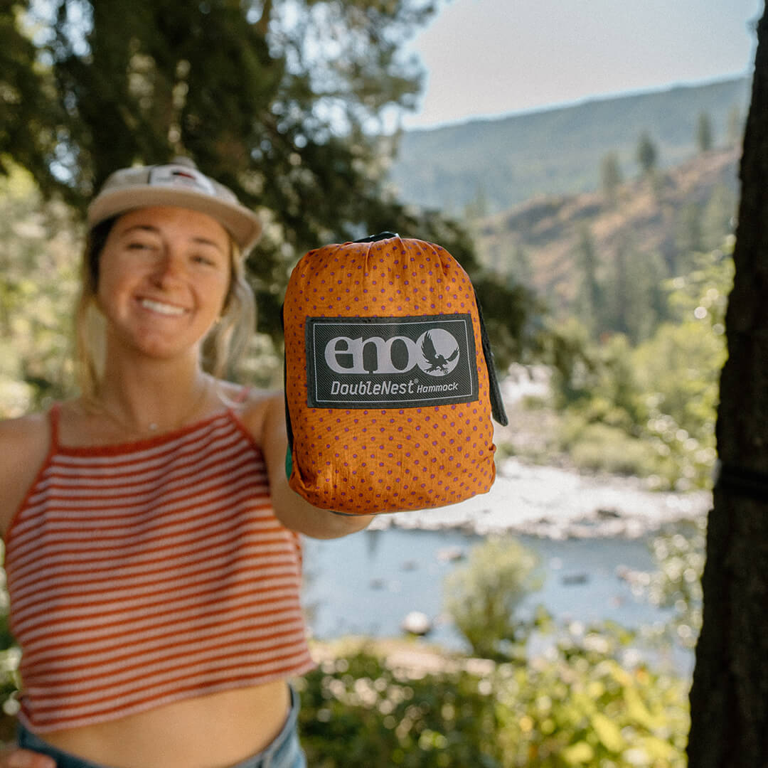 Young woman holding packed ENO DoubleNest Hammock Print stuff sack in front of camera with soft river and forest in the background.