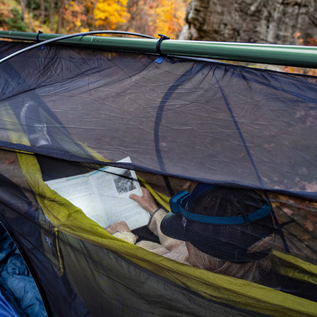 Person lays in hammock inside of the ENO Nomad Shelter System while reading a book with a headlamp.