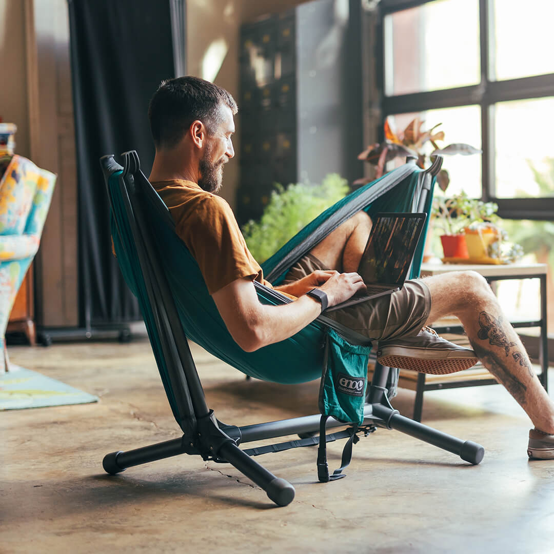 Person sits smiling indoors on an ENO hammock set up on the Parklite Hammock Chair Stand with computer in lap.