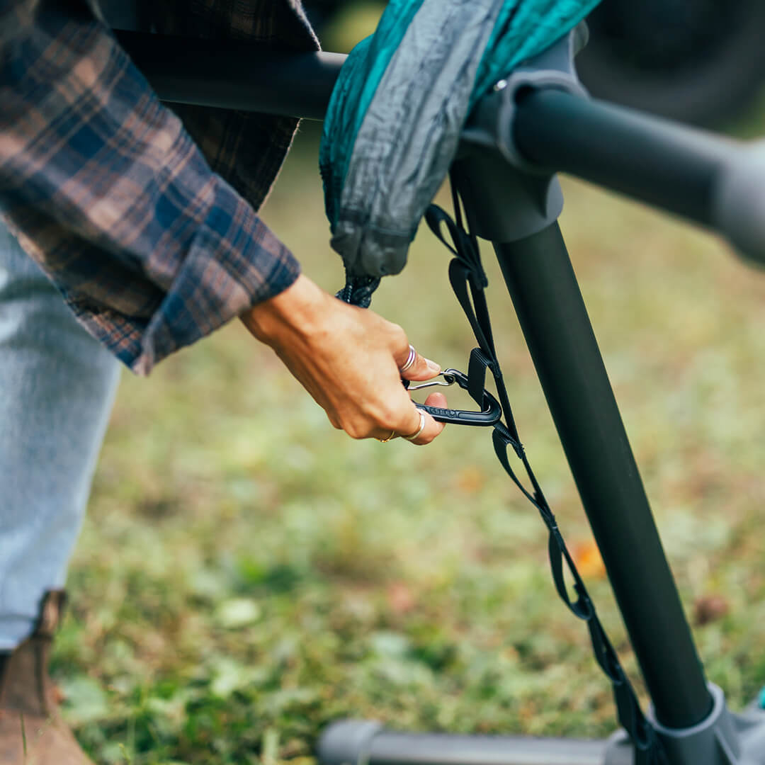 Close up image of hand clipping DoubleNest Hammock carabiner to adjustable strap on the Parklite Hammock Chair stand with blurred background.