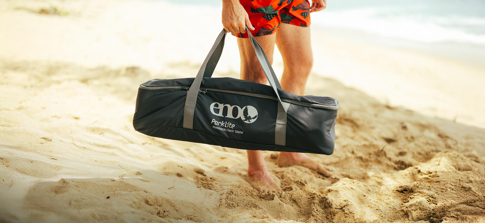 Person holding a Parklite Hammock Chair Stand carrying bag on a sandy beach.