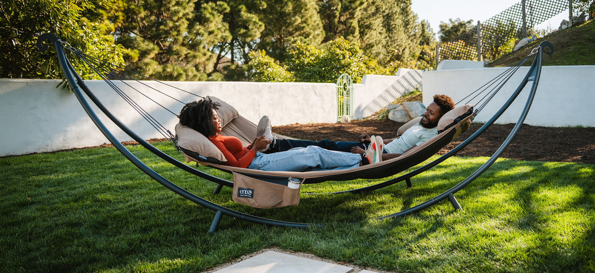 Two people relaxing in a SuperNest Hammock on a SoloPod XL Hammock Stand in a backyard setting.