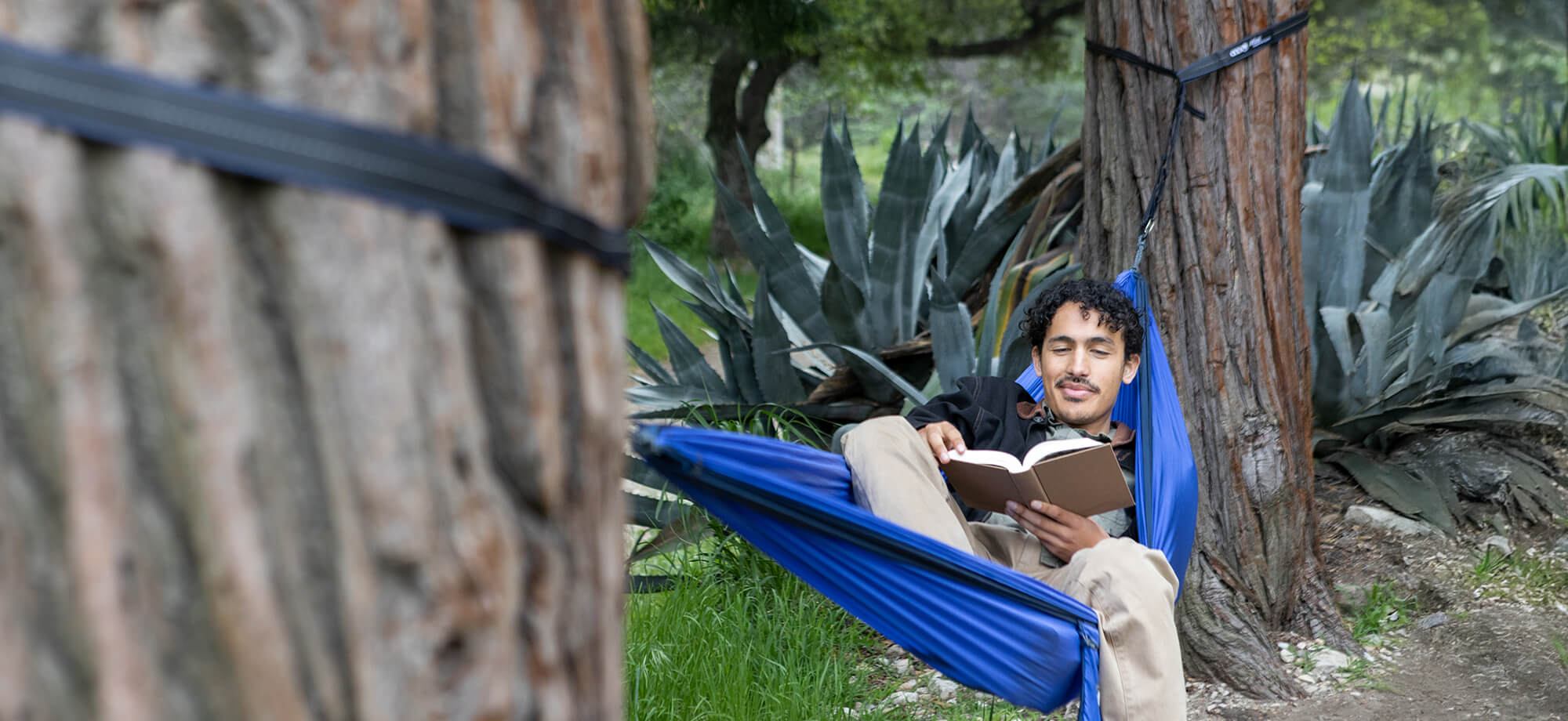 A person lays back while reading a book in an ENO DoubleNest Hammock set up between two trees with the Atlas Hammock Straps.