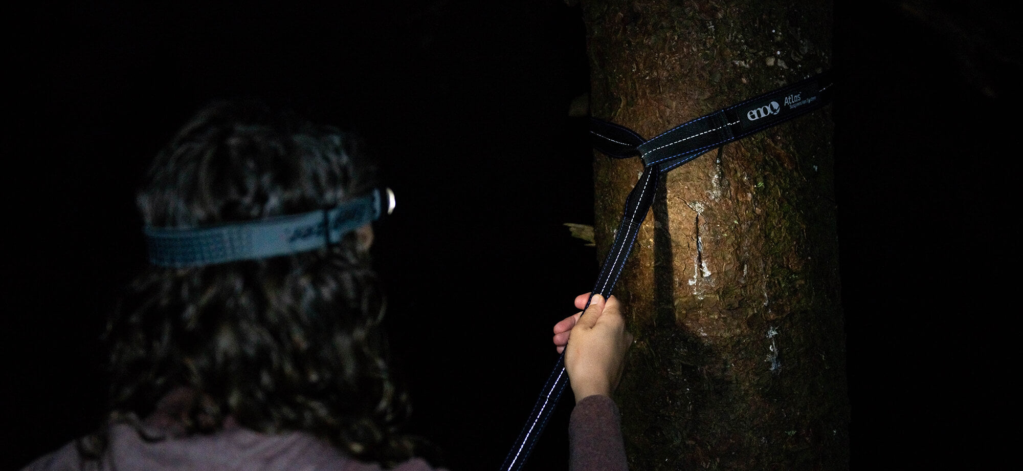 A person set up their ENO Atlas Hammock Straps in the dark with a headlamp illuminating the reflective stitching for nighttime visibility.