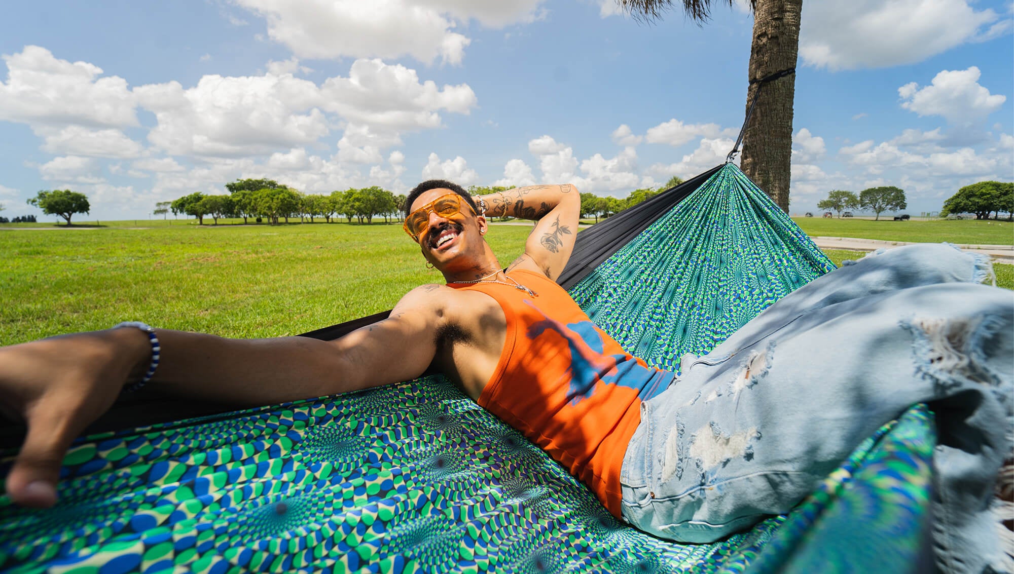 A man wearing a bright orange tank top and distressed light blue jeans relaxes in the ENO DoubleNest Hammock Print in Optical Illusion outdoors under a cloudy sky.