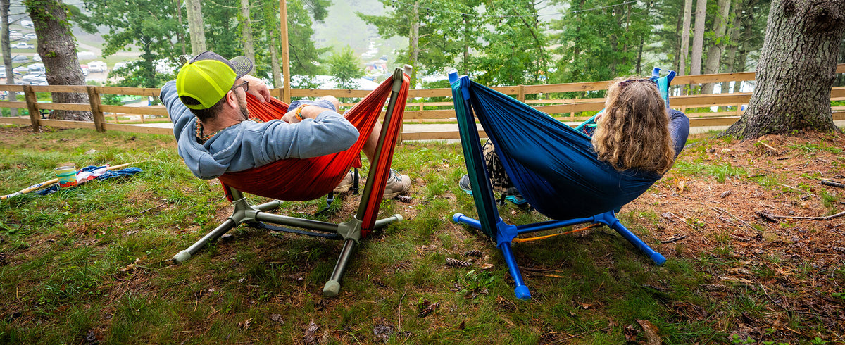 Two people relaxing in ENO Parklite Hammock Chair Stands on a grassy wooded lawn. One person is lying in a red hammock on the left, wearing a bright yellow-green cap and a light blue long-sleeve shirt.