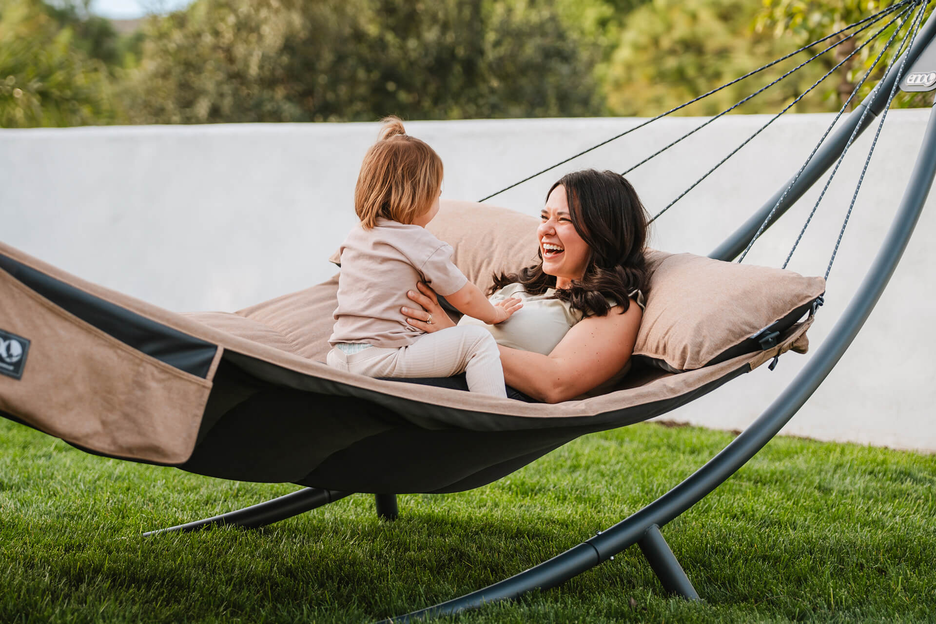 A mother lays in her SuperNest Hammock laughing with their child sitting in their lap on a SoloPod XL Hammock Stand in a backyard.