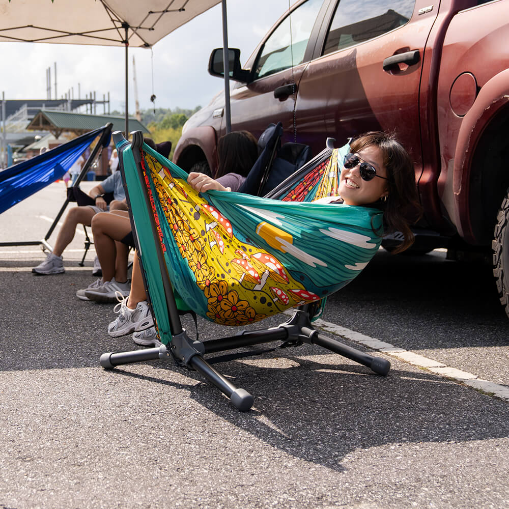 A person sitting back in an ENO DoubleNest Print Hammock set up on a Parklite Hammock Chair Stand in a tailgating setting.