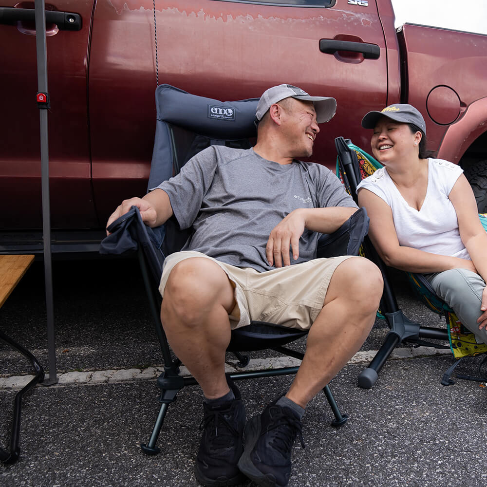 Two people sitting in front of a red truck, one in an ENO Lounger DL Mesh Chair and the other in the Parklite Hammock Chair Stand..