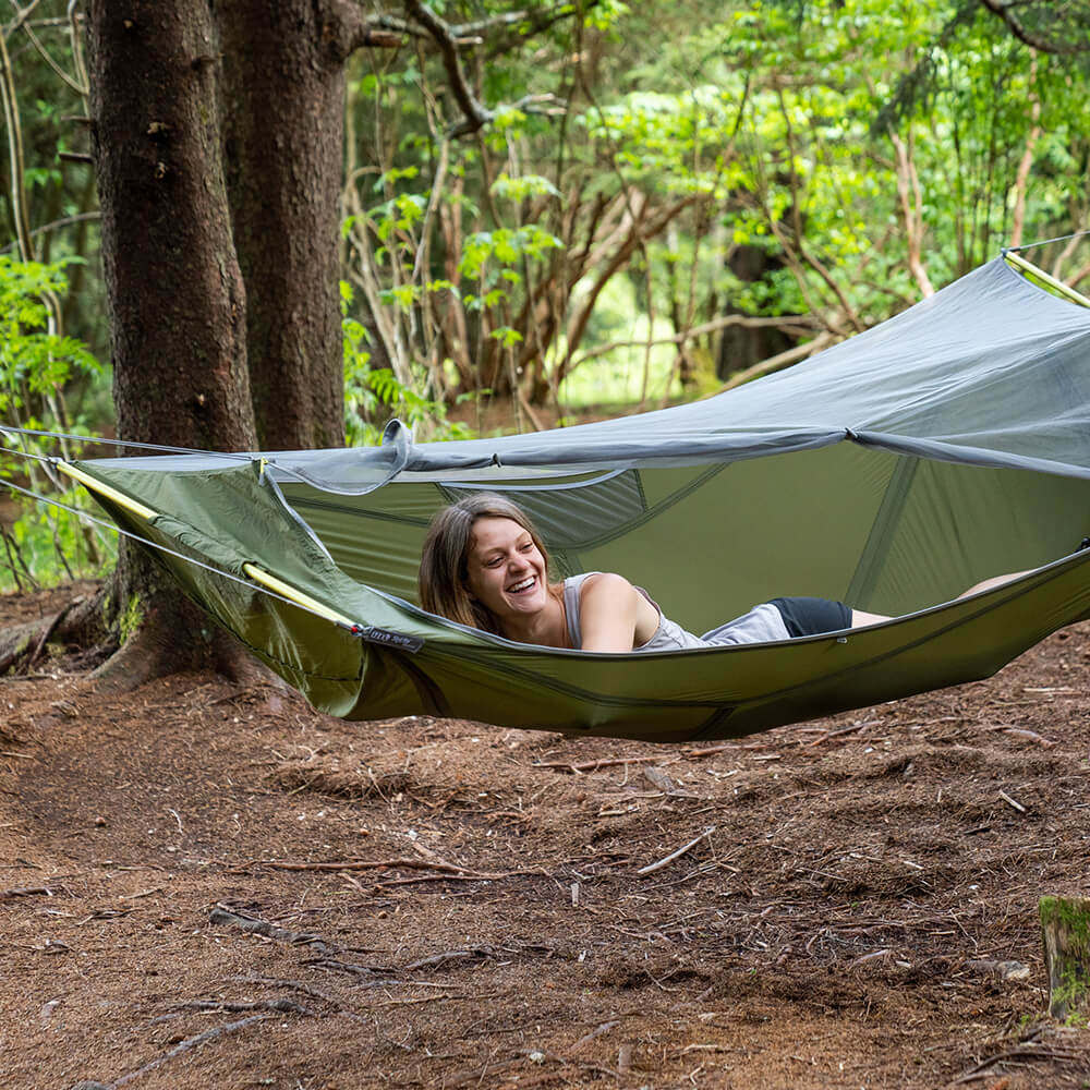 A young smiling woman looks out of a camping hammock in the woods with an included bug net.