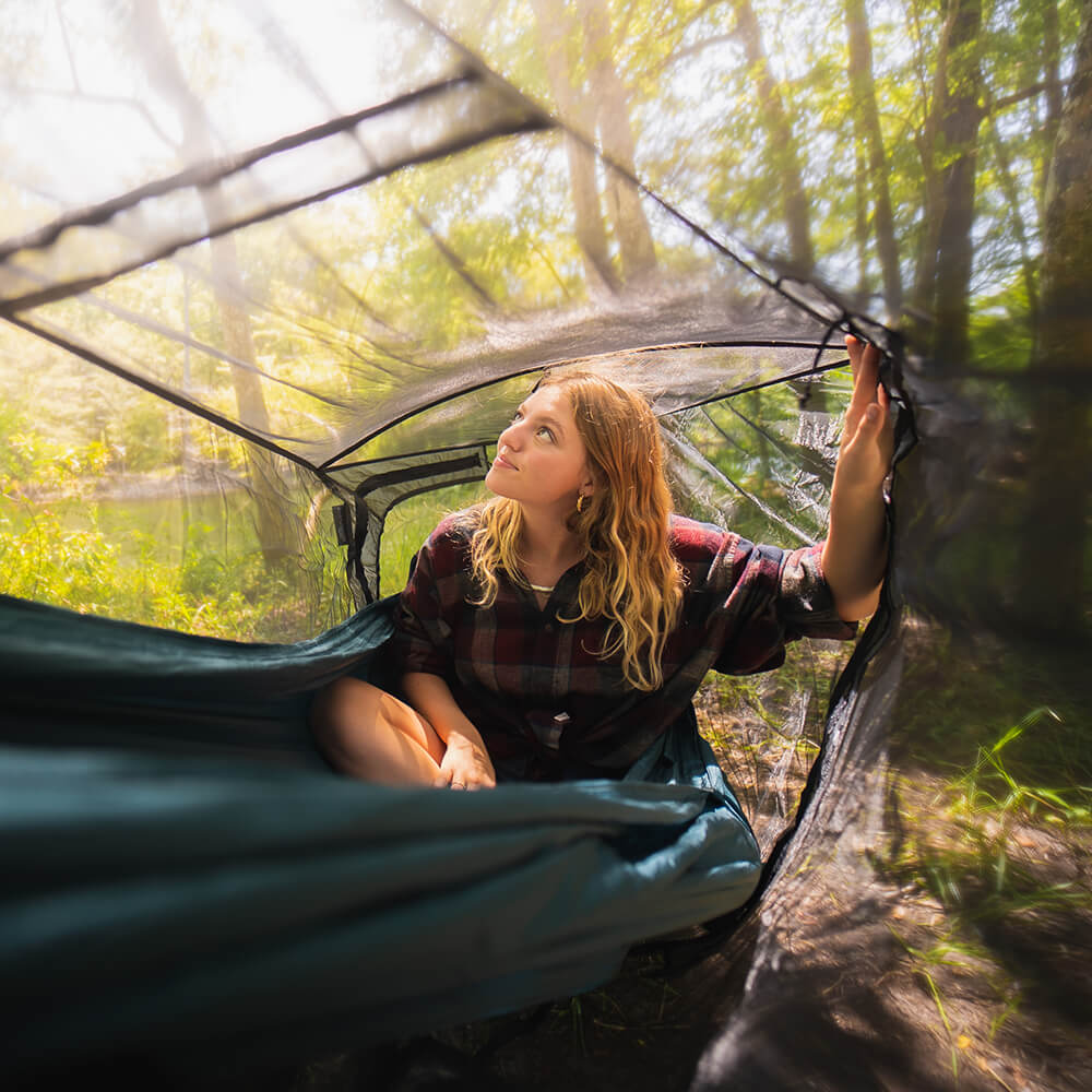 A young woman looks through a bug net into the forest from her portable hammock.