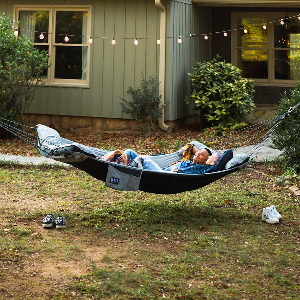 A diverse couple lay in an ENO backyard hammock between two trees in the front yard of a home.