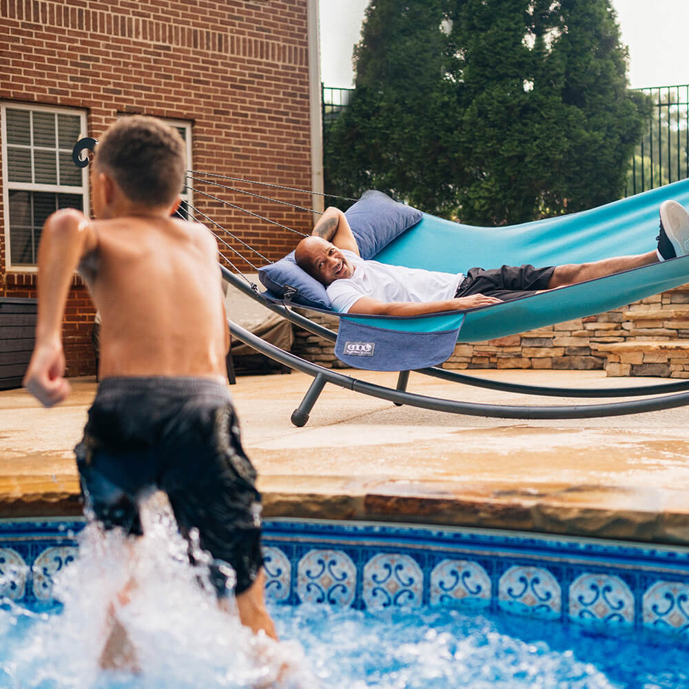 An older diverse man relaxes in an ENO backyard hammock as he watches his child run out of a pool. 