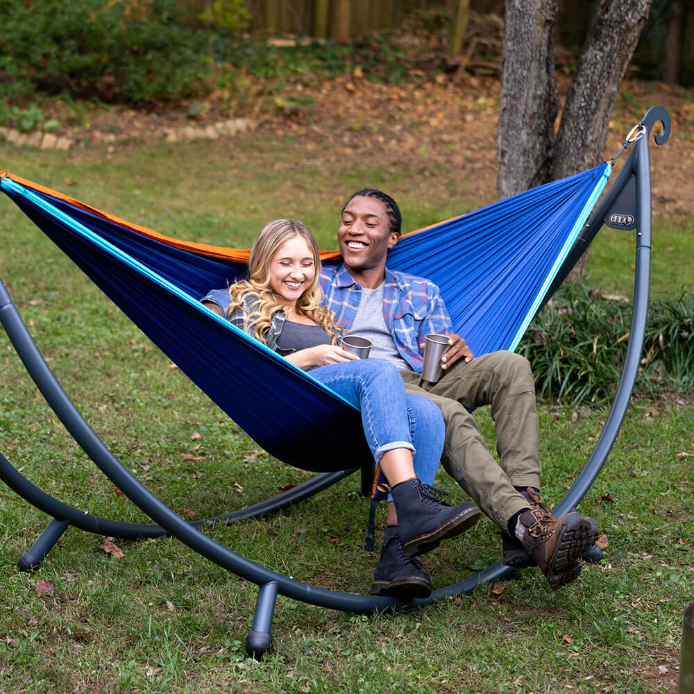 A young diverse couple sits up smiling, relaxed in an ENO portable hammock on a hammock stand in a backyard environment. 