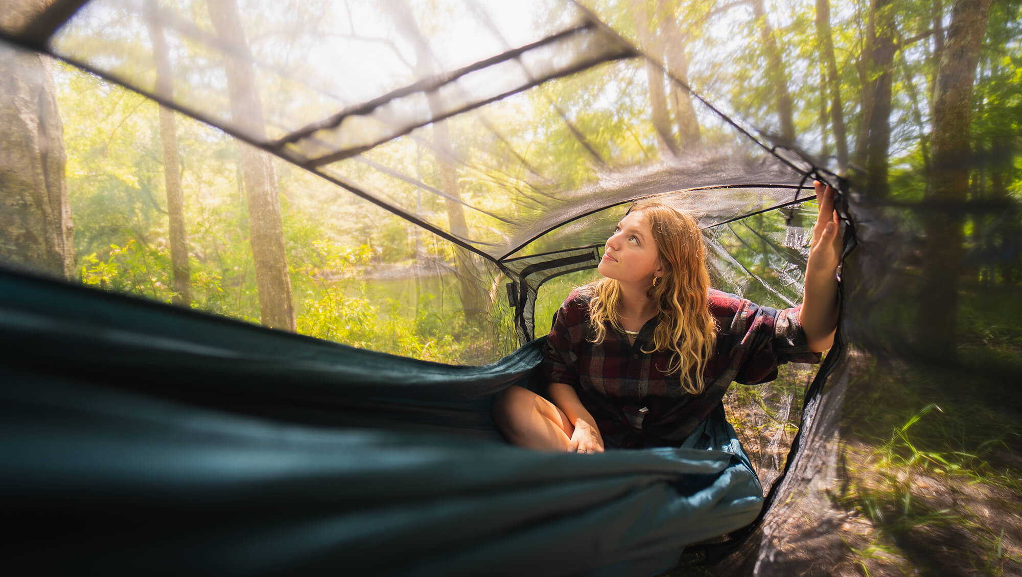 Young woman sits inside ENO Guardian DX Bug Net set up around an ENO Hammock within forest.