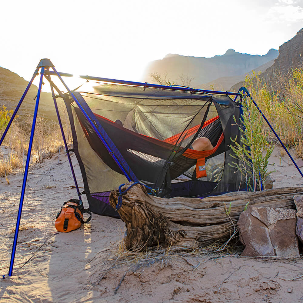 A person lying in an ENO adventure hammock set up on a portable hammock stand in a desert with mountains in the background.