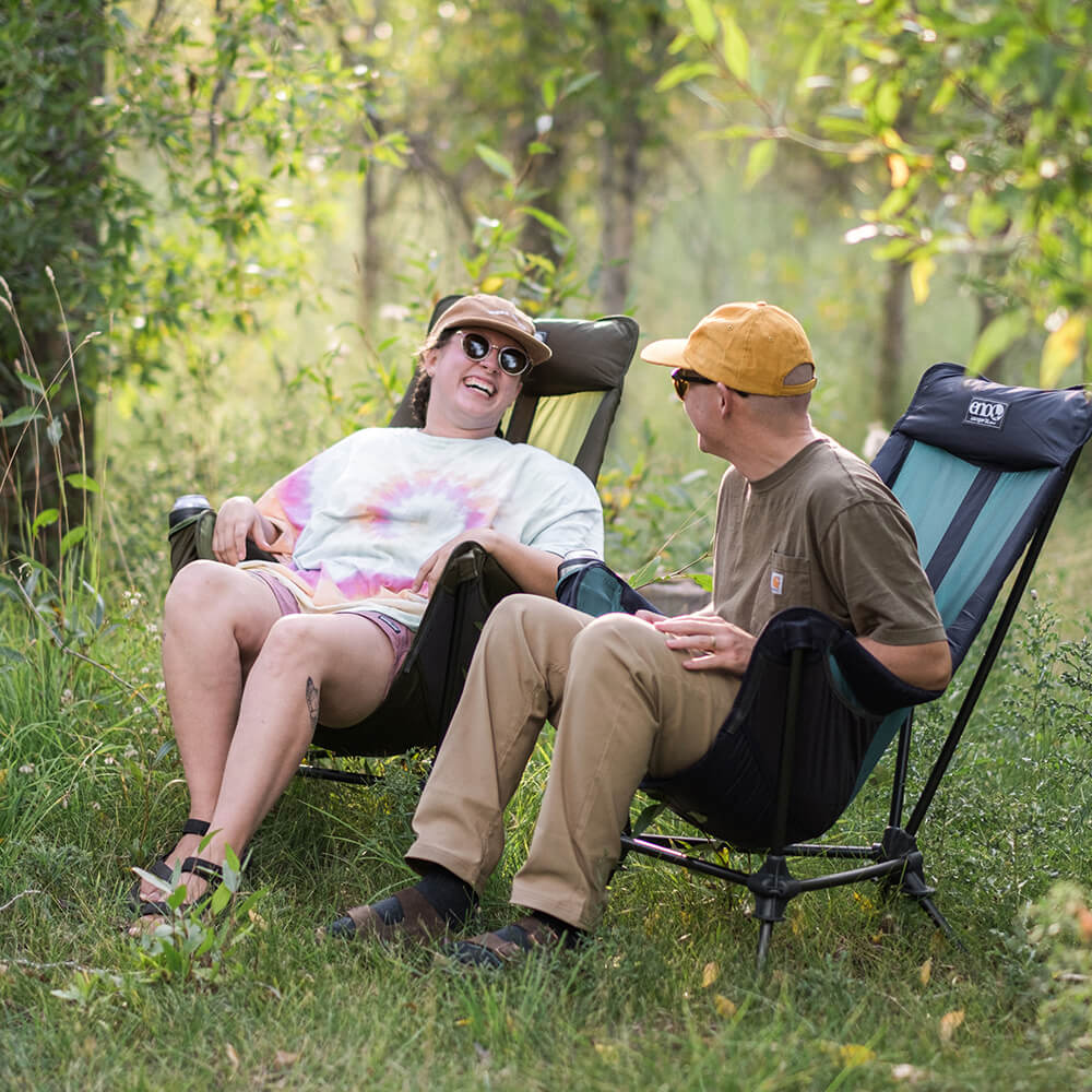 A young couple looks at one another laughing in ENO hammock chairs within lush green landscape. 