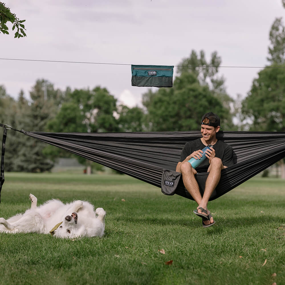 Young man sitting upright in ENO portable hammock watching his dog roll in grass with hammock camping accessories setup above. 