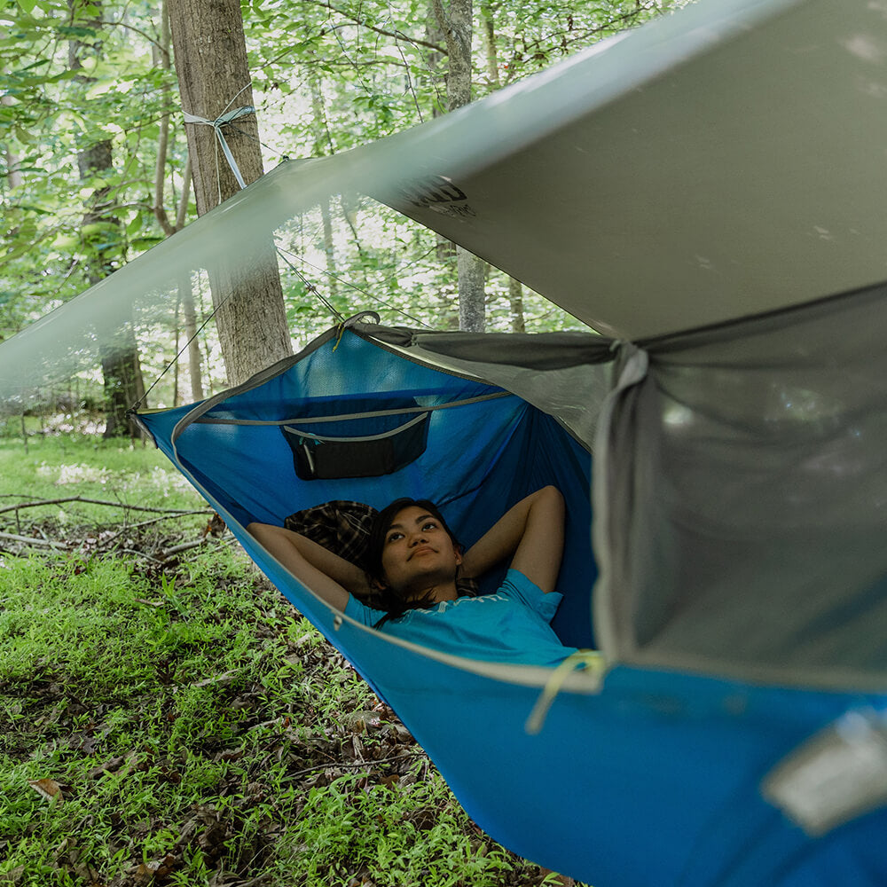 A young woman looks up as she lays back in an ENO camping hammock with a rain tarp setup above in the forest. 