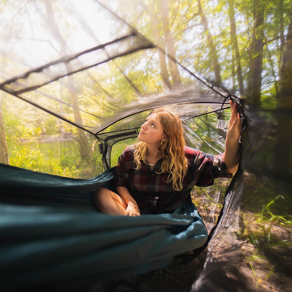 A young woman looks out into the trees in an ENO packable hammock with a hammock bug net.