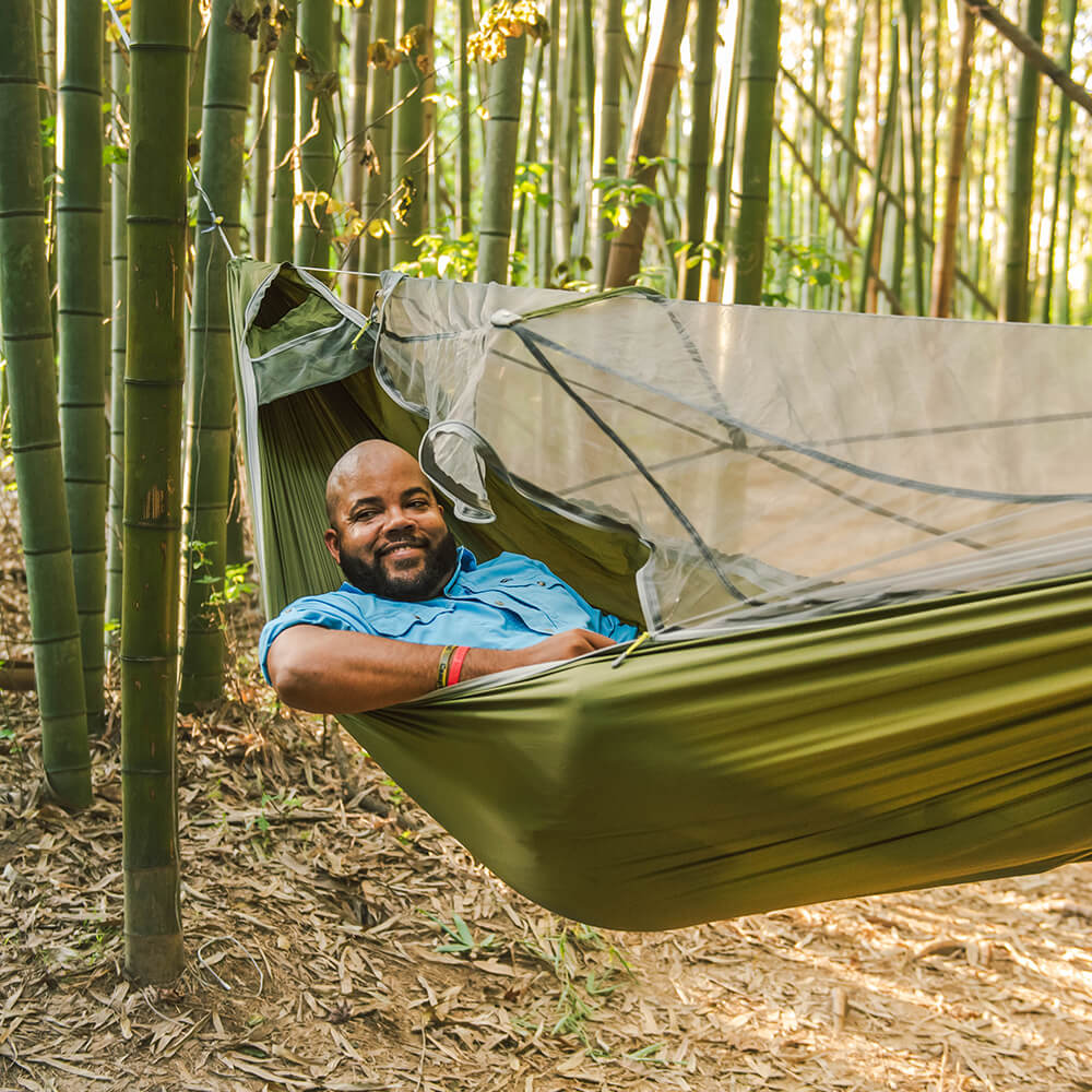 A diverse middle-aged man looks off-camera in a bamboo forest while relaxing in an ENO hammock with an attached bug net.