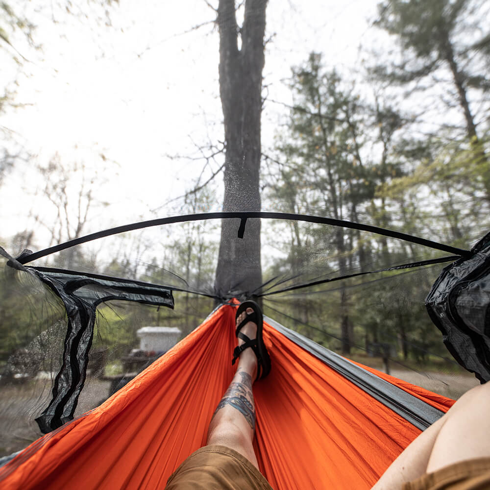 A person's lower body relaxes in an ENO hammock inside of a bug net at a campsite looking up at the ski. 