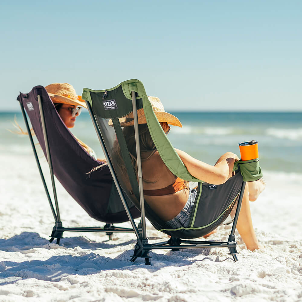 Two women relaxing beachside under blue ski in their ENO hammock chair.