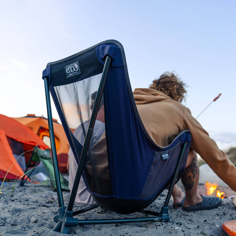 Young man sitting fireside at campsite in ENO hammock camping chair while roasting food.