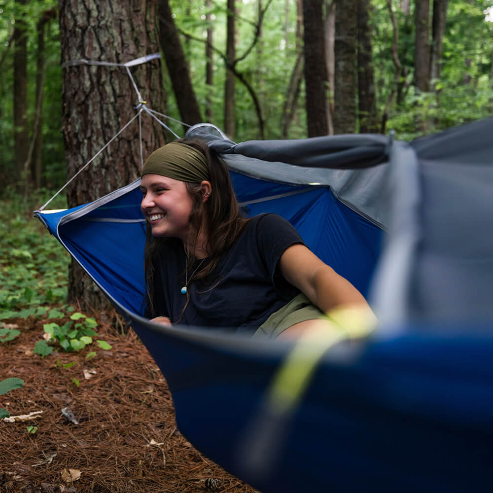 A young woman smiles while relaxing in an ENO portable hammock while camping in the woods.