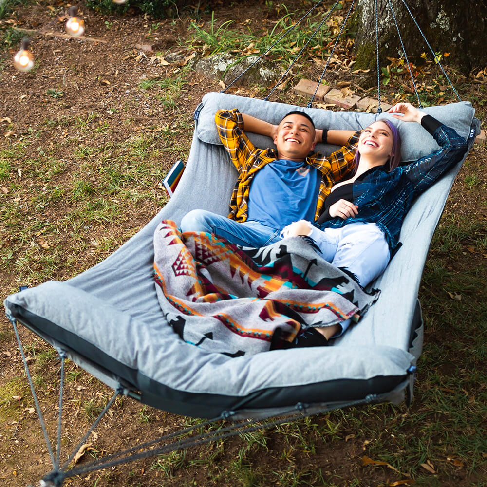 A young diverse couple smiles as they lie down together looking up in an ENO backyard hammock.