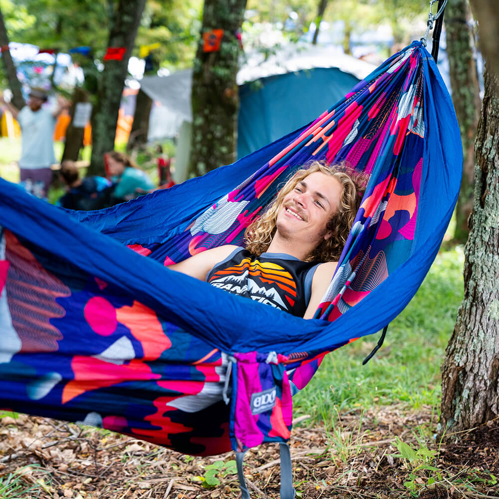 A man rests smiling in an ENO festival hammock off of a tree with tents in the background. 