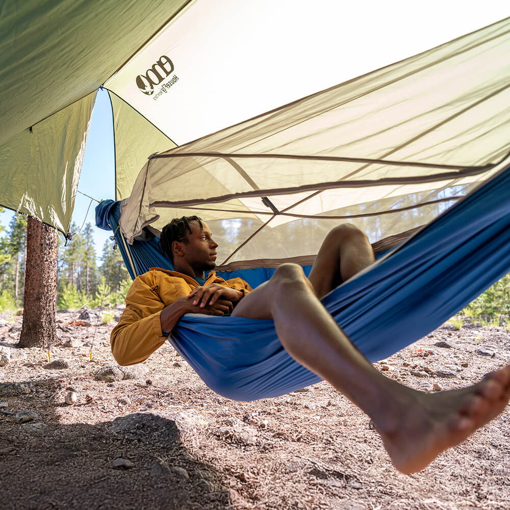 A person laying in an ENO JungleNest Hammock in the wooded backcountry with a HouseFly Rain Tarp set up overhead.