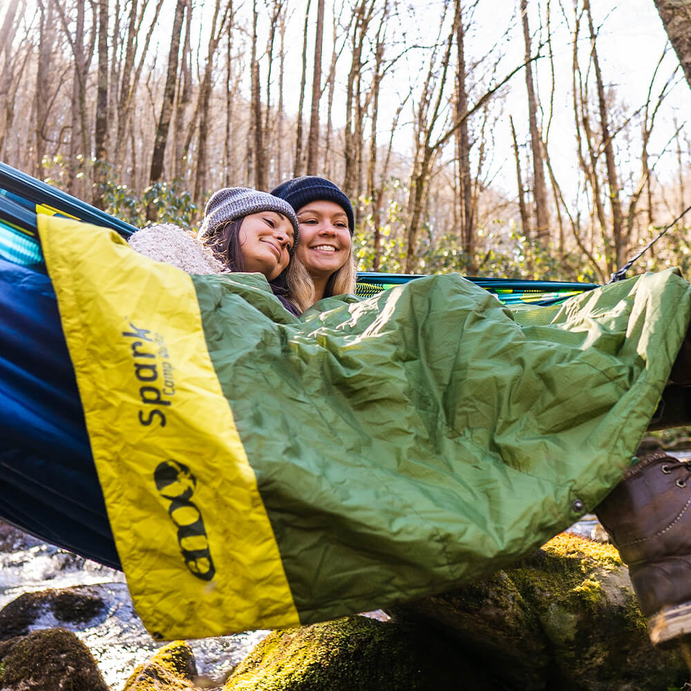 Two women sit smiling in an ENO portable hammock with hammock insulation creekside during winter.