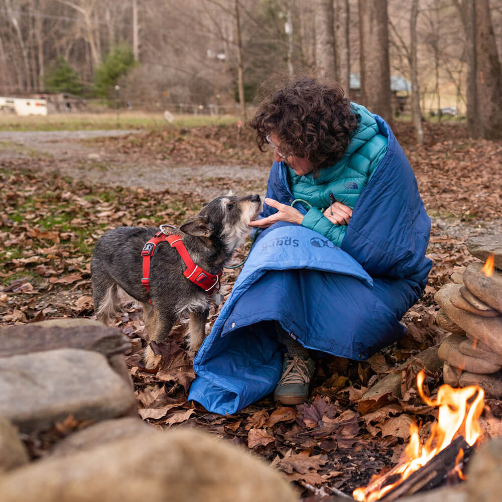 Woman sits fireside at campground wrapped in an ENO camp quilt as she pets little dog on the chin.