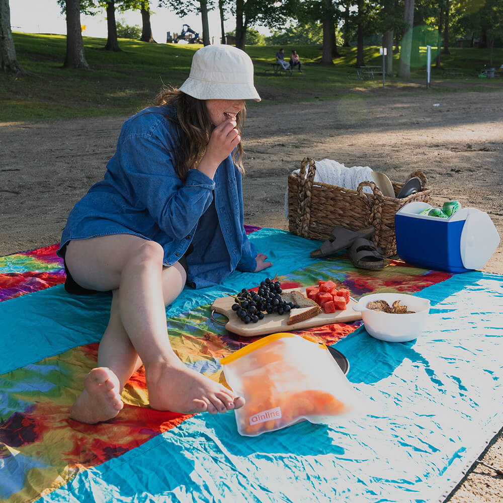 A woman sits on an ENO outdoor picnic blanket while snacking on a fruit bored in the park.