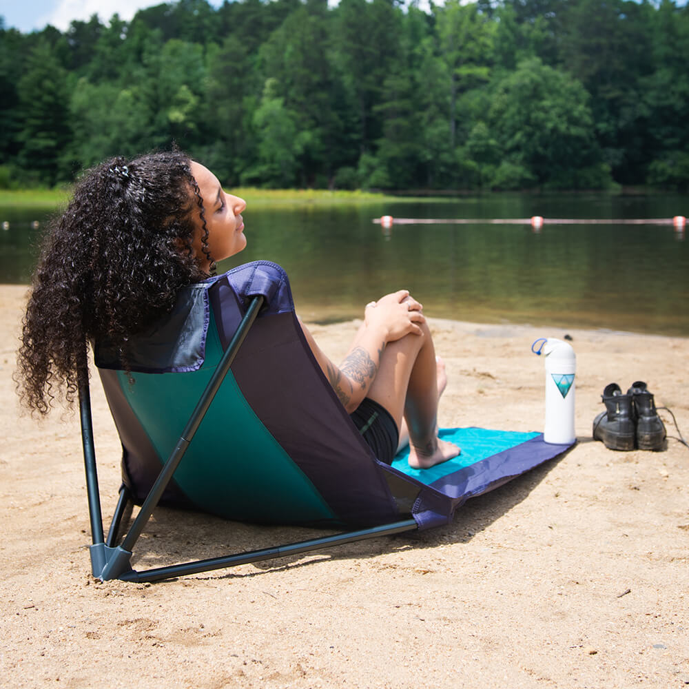 A woman lay relaxing lakeside on an ENO ground chair with shoes off and a water bottle within reach.