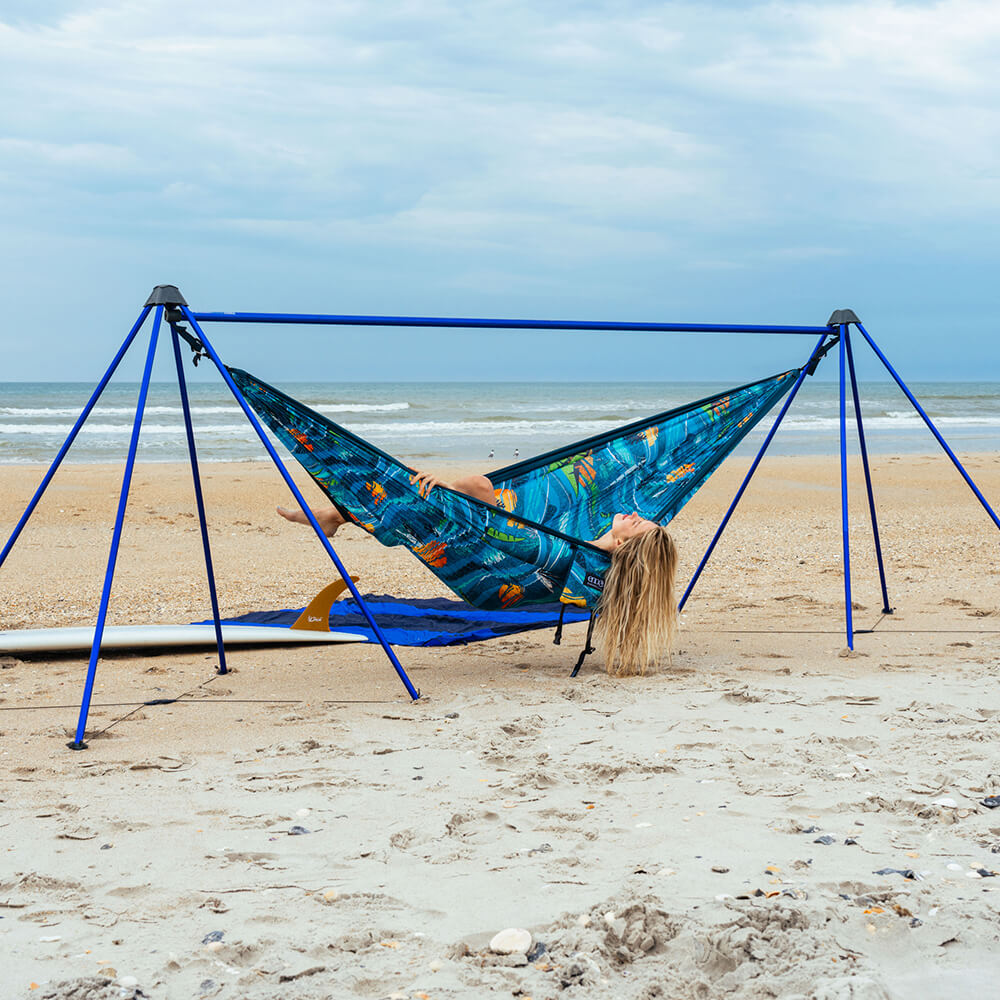 A woman lays back under a blue ski on the beach in an ENO portable hammock stand on the sand.