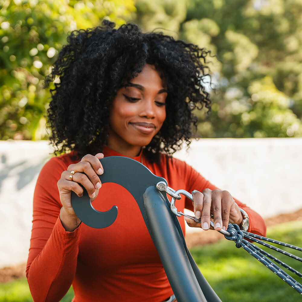 A woman attaching a steel ENO hammock carrabiner to an ENO hammock stand with a blurred green outdoor background