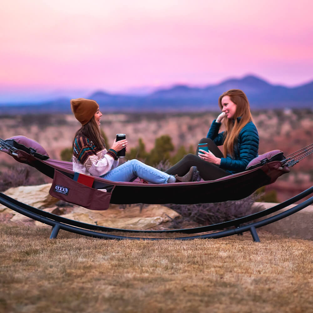 Two women sit facing each other with beverages in an ENO backyard hammock and hammock stand under a pink sunset.