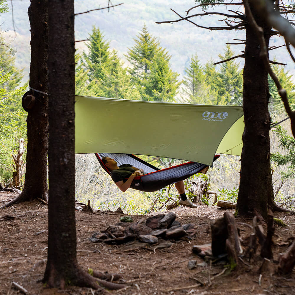 ENO camping tarp setup overtop of ultralight hammock with a young man laying back and legs hanging off in front of a wooded mountain backdrop.