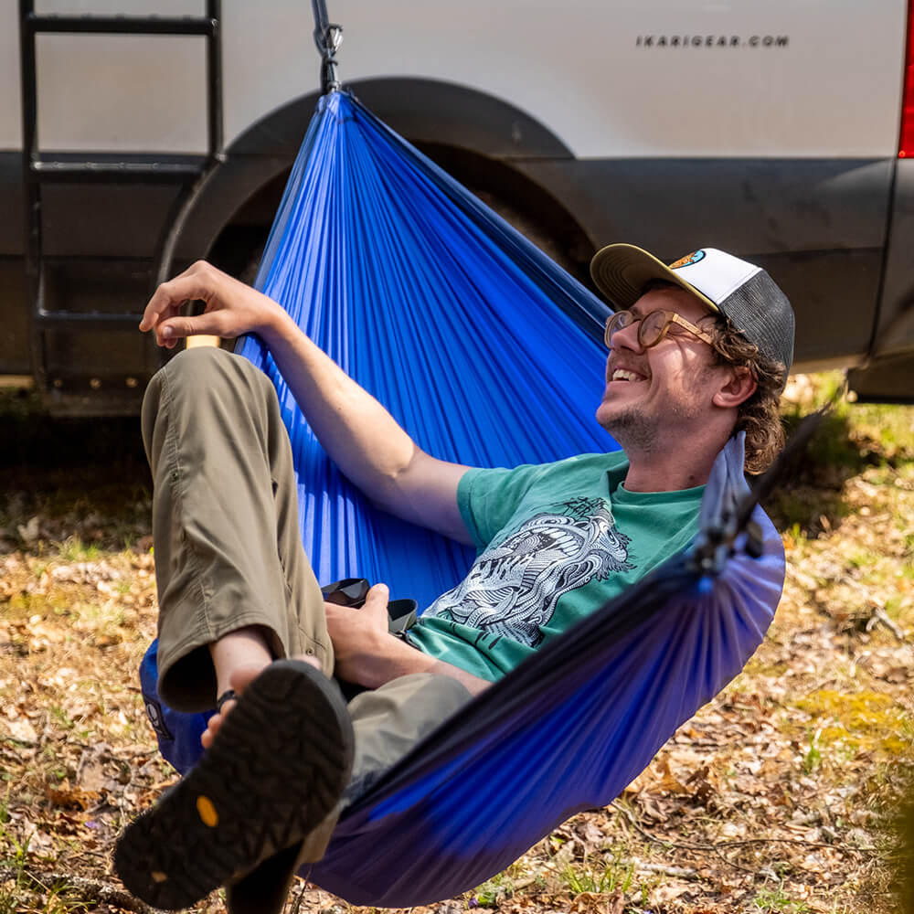 Person sitting back smiling with their feet up in a blue ENO portable hammock with camper can in background.